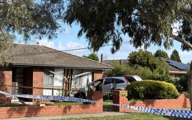 Car crashed into front of brick house, damaging window and wall; police tape cordons off yard in a suburban street.