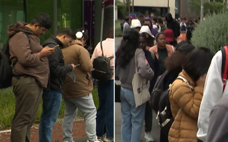 Group of people lined up outdoors, many looking at their smartphones while waiting outside a building.
