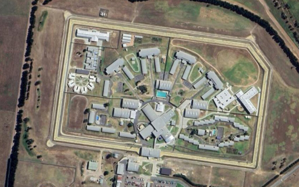 Aerial view of a large, high-security prison with interconnected buildings inside a fenced perimeter and surrounding fields.