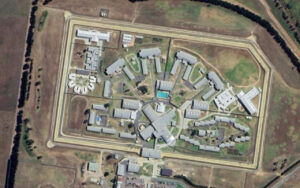 Aerial view of a large, high-security prison with interconnected buildings inside a fenced perimeter and surrounding fields.