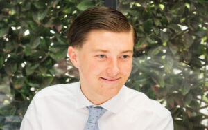 Smiling young man in a white dress shirt and light blue polka-dot tie, head-and-shoulders portrait with a leafy background.