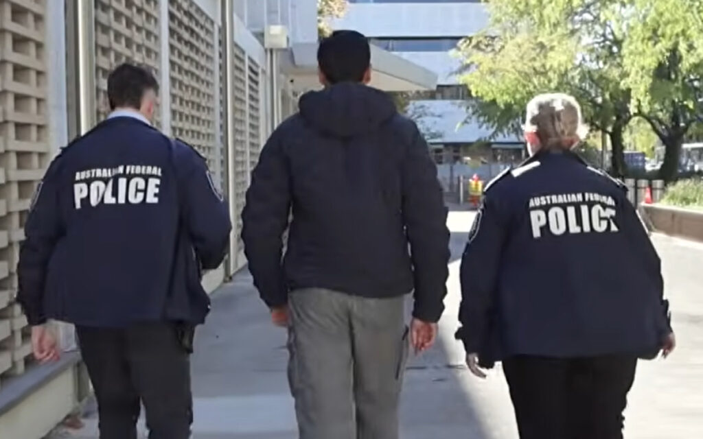 Two Australian Federal Police officers walk on either side of a civilian, escorting him down a sidewalk.