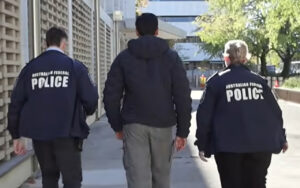 Two Australian Federal Police officers walk on either side of a civilian, escorting him down a sidewalk.
