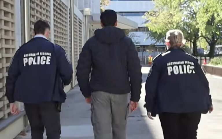 Two Australian Federal Police officers walk on either side of a civilian, escorting him down a sidewalk.