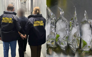 Two Australian Border Force officers escort a detainee down a hallway, jackets reading AUSTRALIAN BORDER FORCE, detainee in custody.