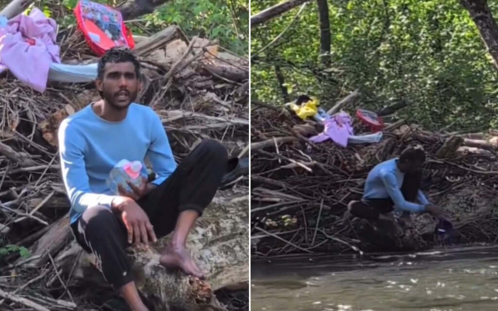 Man in a light blue sweater sits on a log amid a pile of branches and clothes by a muddy riverbank, holding a plastic bottle.