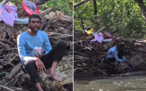 Man in a light blue sweater sits on a log amid a pile of branches and clothes by a muddy riverbank, holding a plastic bottle.