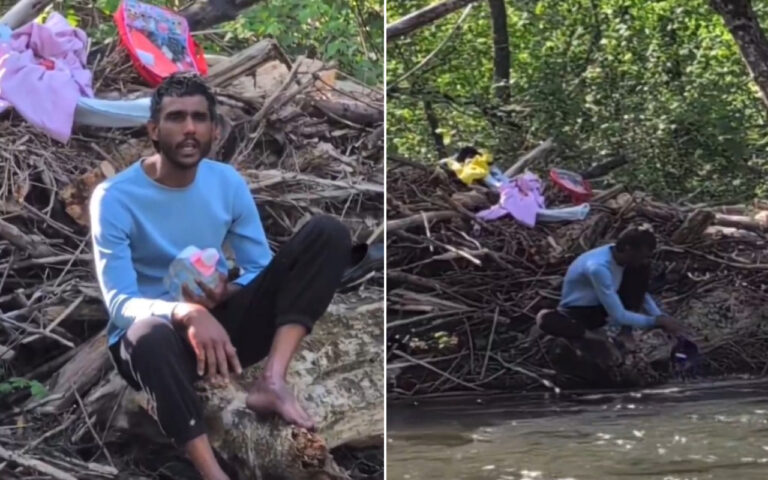 Man in a light blue sweater sits on a log amid a pile of branches and clothes by a muddy riverbank, holding a plastic bottle.