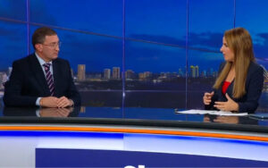 Two news anchors sit at a glass desk in a studio, speaking across from each other against a city skyline backdrop.