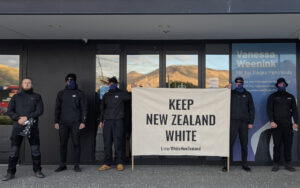 Six men in black jackets stand outside a storefront, holding a beige banner reading 'KEEP NEW ZEALAND WHITE'.