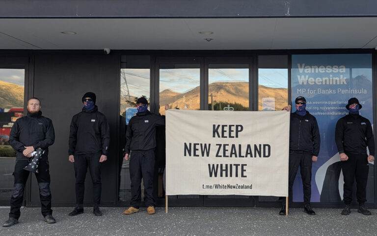 Six men in black jackets stand outside a storefront, holding a beige banner reading 'KEEP NEW ZEALAND WHITE'.