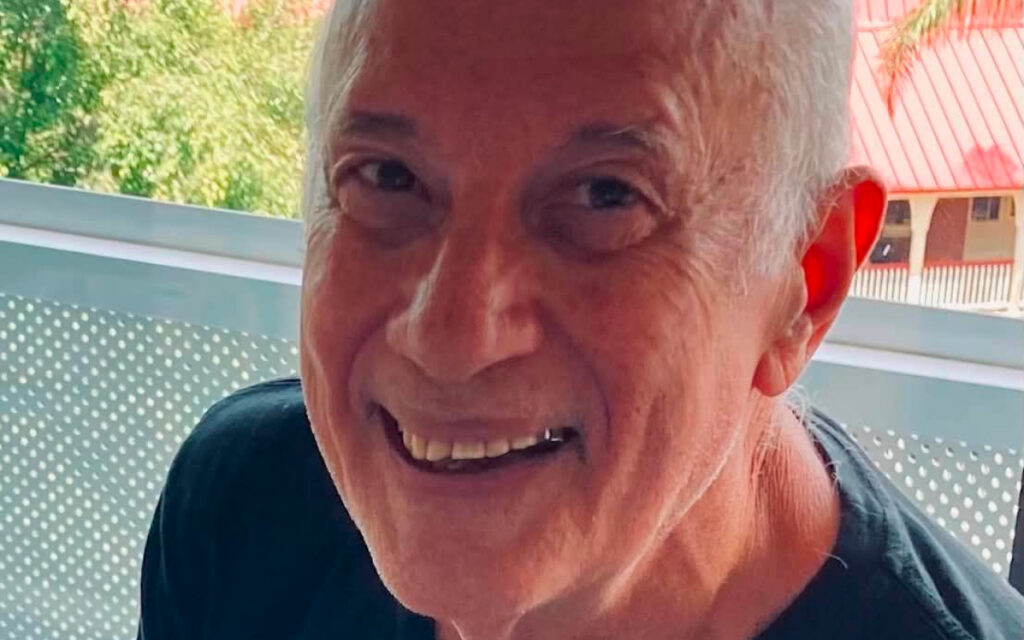 Close-up of an older man smiling outdoors, with short gray hair and a dark shirt, seen from the shoulders up.