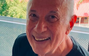 Close-up of an older man smiling outdoors, with short gray hair and a dark shirt, seen from the shoulders up.