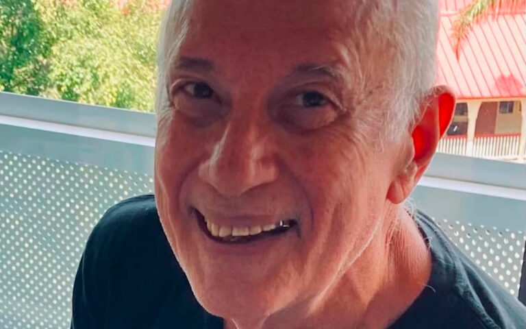 Close-up of an older man smiling outdoors, with short gray hair and a dark shirt, seen from the shoulders up.