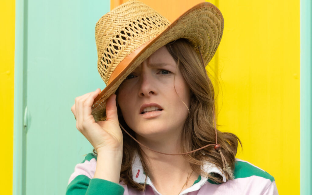 Woman wearing a wide straw hat, adjusting it with her hand in front of a bright yellow and teal background (outdoor scene).