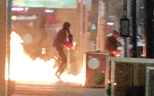 Person in a dark jacket running past a large outdoor flame on a city street at night, with structures and bystanders in the background.