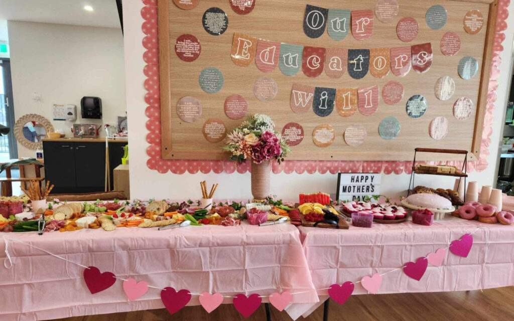 Buffet table for a Mother's Day event, covered in pink tablecloth with a heart garland, offering pretzels, bread, fruit, donuts, pastries, and a vase of flowers under a decorative bulletin board.