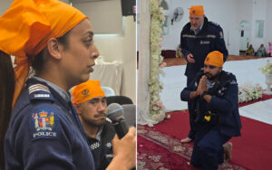 Side-profile of a female police officer in an orange headscarf speaking into a microphone at an indoor event with a seated colleague nearby