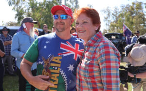 Two people pose for a photo at an outdoor gathering: a man in a Union Jack/Australia-themed shirt and red cap with sunglasses, and a woman in a red-blue plaid shirt smiling beside him, with a crowd and cameras in the background.