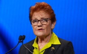 Older woman with short red hair and glasses speaking at a microphone on a blue backdrop, wearing a lime green shirt and pin on a pinstripe blazer