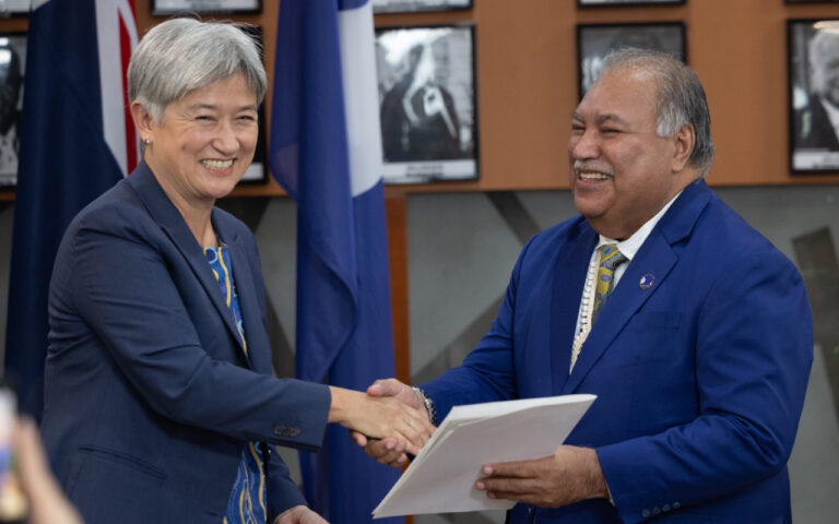 Two professionals in blue suits shake hands during a formal ceremony, one holding a folder.
