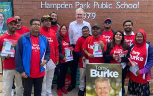 Group of volunteers in red Tony Burke shirts pose outside Hampden Park Public School for a campaign photo. Neck signs and flyers visible.