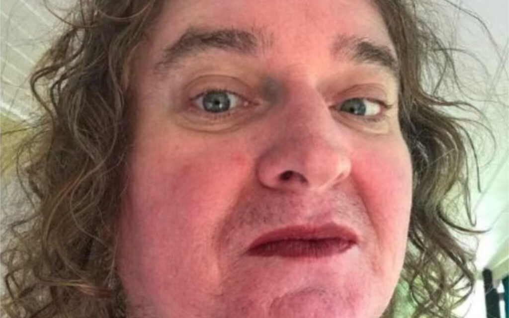 Close-up selfie of a man with messy, shoulder-length hair, stubble, and a surprised, half-smile expression looking at the camera.