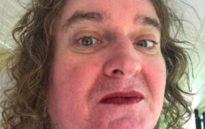 Close-up selfie of a man with messy, shoulder-length hair, stubble, and a surprised, half-smile expression looking at the camera.