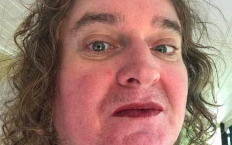 Close-up selfie of a man with messy, shoulder-length hair, stubble, and a surprised, half-smile expression looking at the camera.