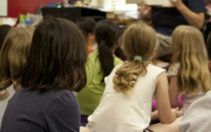 Group of children seated on the classroom floor, facing a teacher who is presenting at the front of the room.