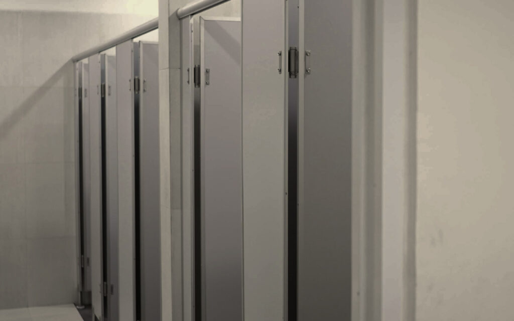 Row of toilet stalls in a public restroom with gray metal doors along a tiled wall, viewed from the hallway.