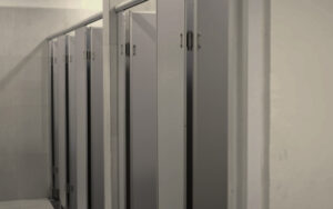 Row of toilet stalls in a public restroom with gray metal doors along a tiled wall, viewed from the hallway.