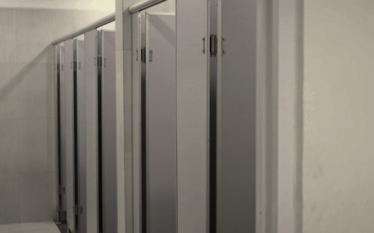 Row of toilet stalls in a public restroom with gray metal doors along a tiled wall, viewed from the hallway.