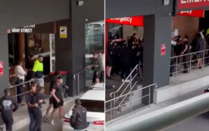 People gathered and lining up outside a building with an emergency entrance sign and a railing along the sidewalk.