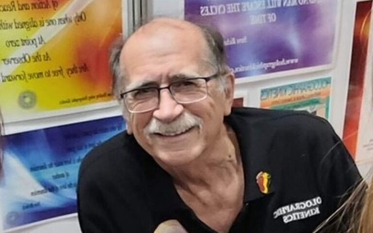 Smiling older man with glasses and a mustache, wearing a black polo shirt with a small colorful pin, in front of vibrant posters