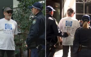 Two men in white anti-Semitism shirts talk with police officers on a city street; one man wears sunglasses and a cap.