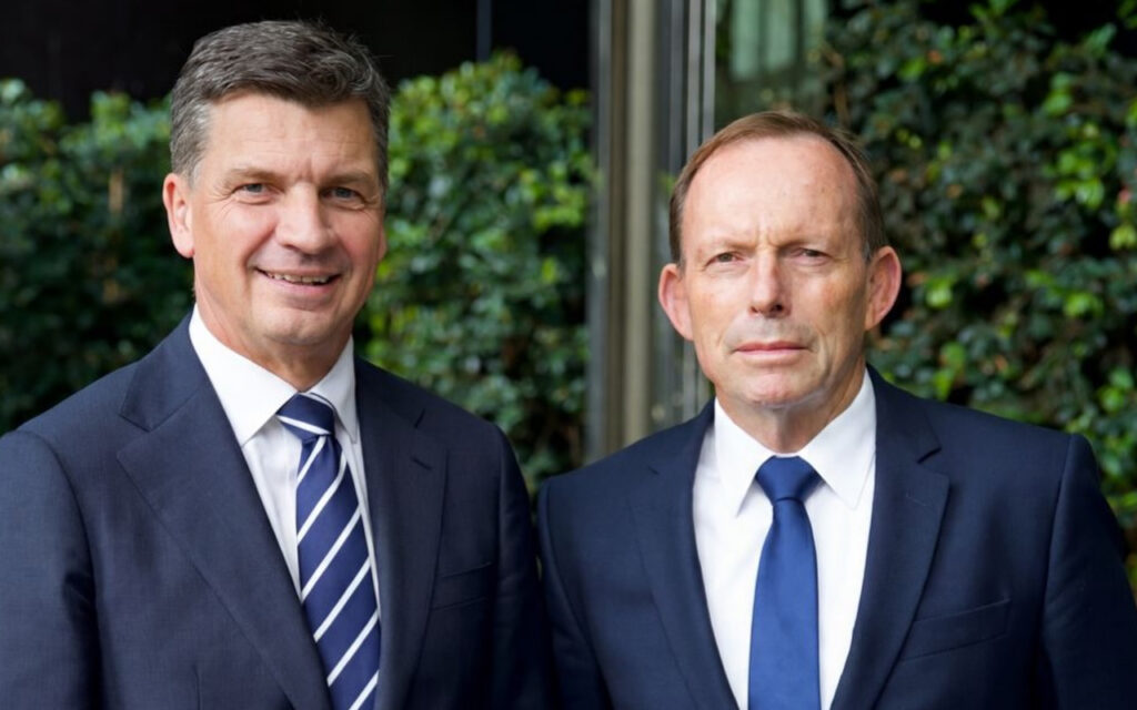 Two businessmen in dark suits pose outside, smiling side by side with greenery behind them.