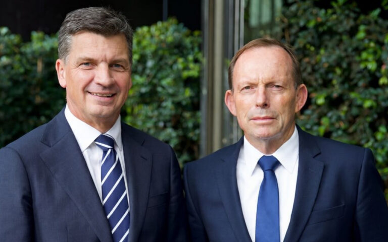 Two businessmen in dark suits pose outside, smiling side by side with greenery behind them.