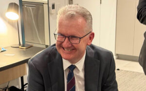 Smiling man in a suit and glasses seated in a modern office/kitchen area.