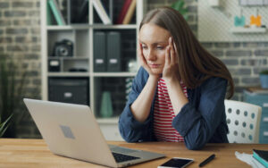 Woman sits at a wooden desk with her head in her hands, looking stressed while using a silver laptop in a modern workspace.