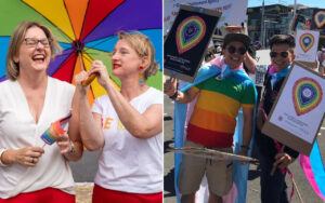 Two women laughing under a large multicolored rainbow umbrella; one holds a small rainbow flag.