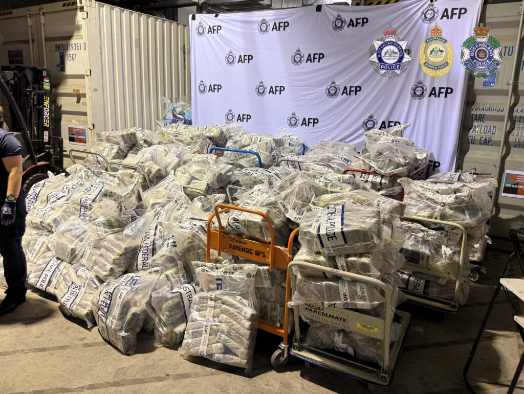 Row of white body bags labeled AFP and forensic stacked in a warehouse beside a police banner backdrop.