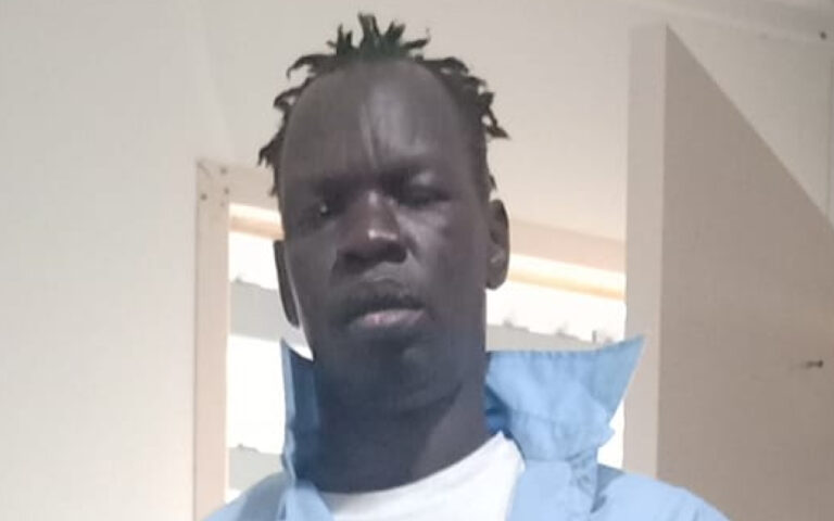 Young man with dark skin wearing a light blue jacket and white shirt, standing indoors near an open doorway.