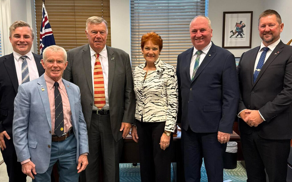 Six adults in business attire posing for a group photo in a meeting room, with a flag in the background and blinds/windows behind them.