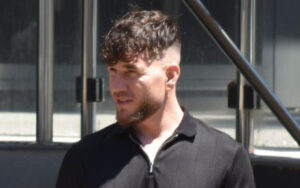 Side-profile of a man with dark hair and stubble wearing a black zip-up polo, outdoors near glass railings.