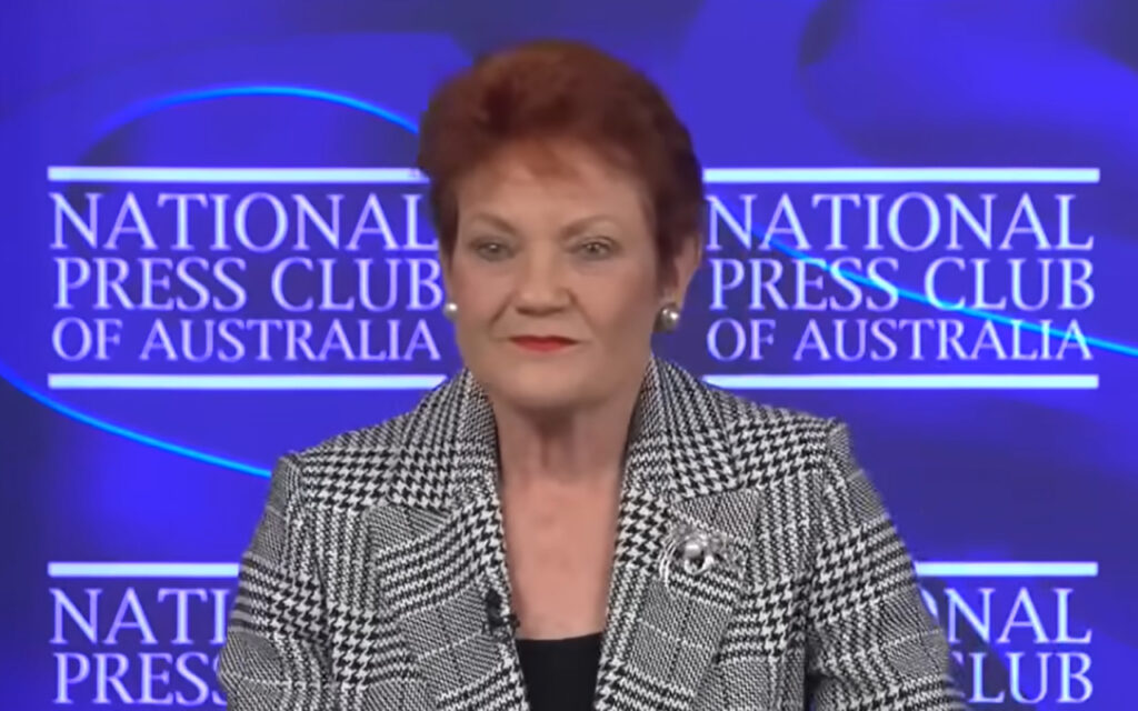 Woman in a black-and-white houndstooth blazer speaks at the National Press Club of Australia, with a lapel microphone visible on the jacket.