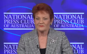 Woman in a black-and-white houndstooth blazer speaks at the National Press Club of Australia, with a lapel microphone visible on the jacket.