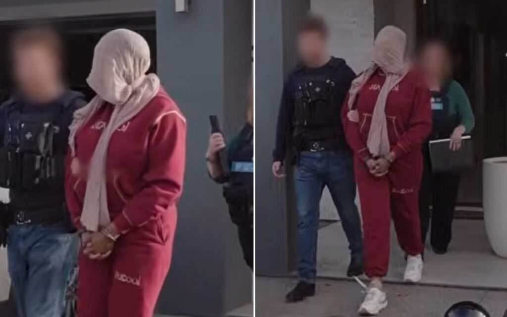 Suspect in a red tracksuit with a scarf over the head, handcuffed, escorted by officers outside a building.”],