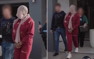 Suspect in a red tracksuit with a scarf over the head, handcuffed, escorted by officers outside a building.”],