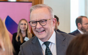 Smiling older man in a gray suit and black glasses at a networking event, with a pink banner in the background.